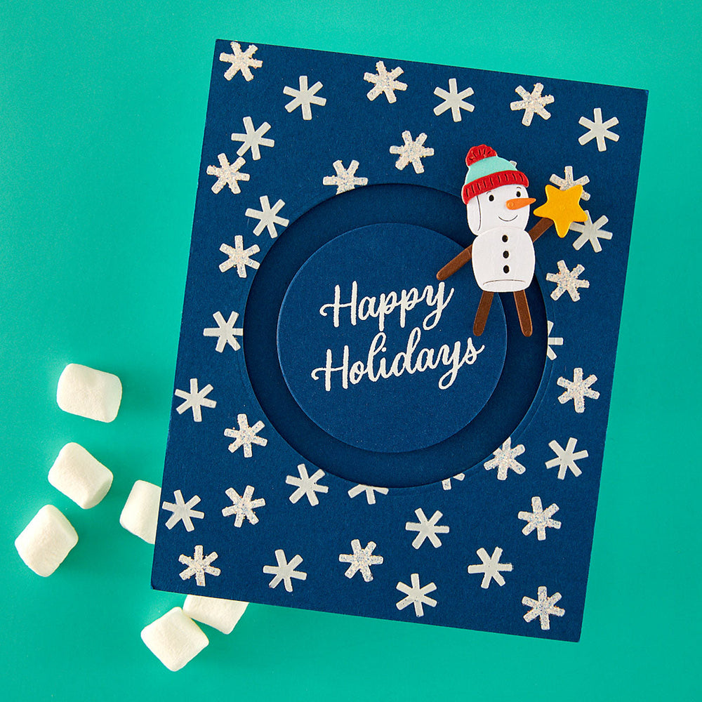 A square navy blue handmade holiday card with a circular cutout in the center and the words 'Happy Holidays' in white script. The card is decorated with white snowflakes and features a paper snowman wearing a red and green hat and brown arms, holding a yellow star.