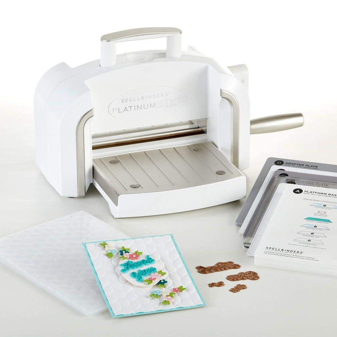 A Platinum SIX die cutting and embossing machine with a Universal Plate System. The machine is white with brushed platinum trim, next to a crafted card and additional embossing folders.