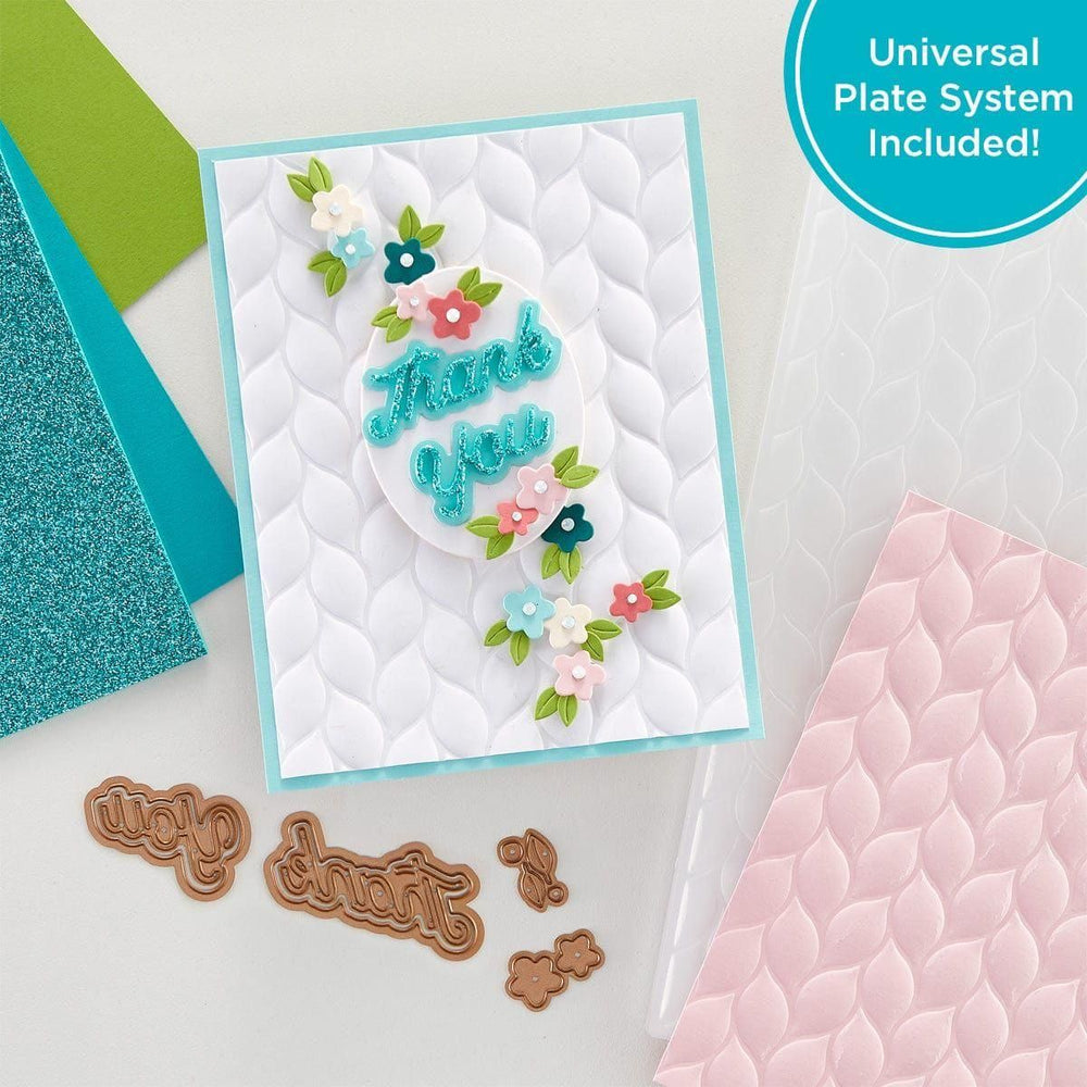 A Platinum SIX die cutting and embossing machine with a Universal Plate System. The machine is white with brushed platinum trim, next to a crafted card and additional embossing folders.